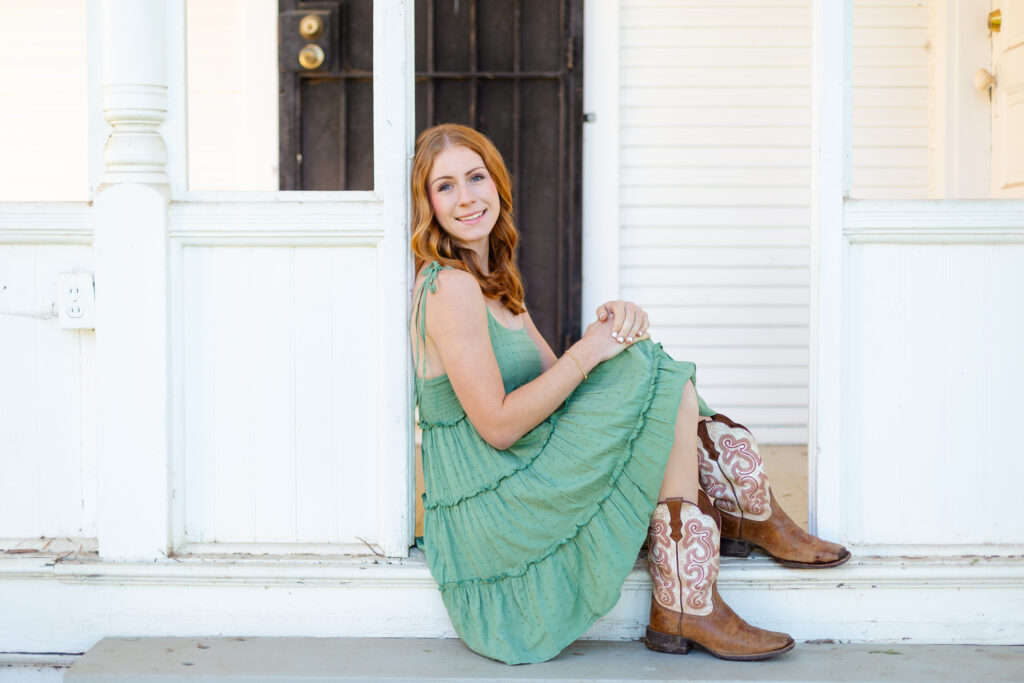 a woman wearing a green dress and cowboy boots sitting on a porch, posing for photography that will be used in blogging.